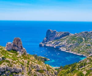 Et fantastisk kystlandskab med klipper og turkisblåt vand. Talrige både ligger for anker i bugten, omgivet af frodig grøn vegetation under en klar blå himmel.