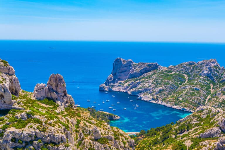 Et fantastisk kystlandskab med klipper og turkisblåt vand. Talrige både ligger for anker i bugten, omgivet af frodig grøn vegetation under en klar blå himmel.