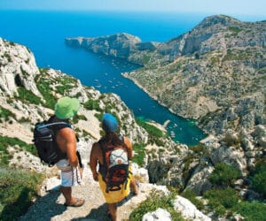 To vandrere med rygsække står på en sti med udsigt over en fantastisk kystudsigt. Landskabet byder på stenede klipper, et klart blåt hav og små både i vandet. Scenen er lys og solrig og fanger naturens skønhed og eventyret.