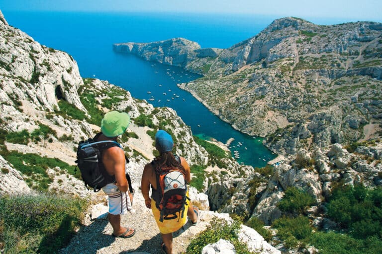 To vandrere med rygsække står på en sti med udsigt over en fantastisk kystudsigt. Landskabet byder på stenede klipper, et klart blåt hav og små både i vandet. Scenen er lys og solrig og fanger naturens skønhed og eventyret.