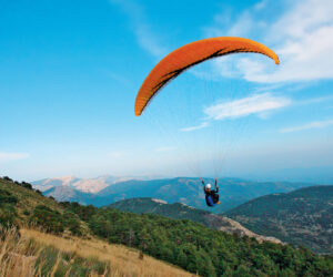 En paraglider med en orange baldakin svæver over et bjergrigt landskab, omgivet af grønne bakker og en flade af blå himmel med et par hvide skyer.
