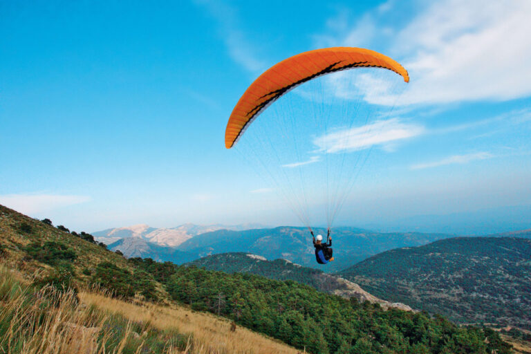 En paraglider med en orange baldakin svæver over et bjergrigt landskab, omgivet af grønne bakker og en flade af blå himmel med et par hvide skyer.