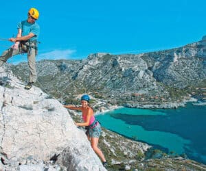 To personer klatrer på en solskinsdag med udsigt over et naturskønt kystlandskab med turkisblåt vand og klipper. Klatrerne bærer hjelme og klatreudstyr, og baggrunden omfatter en bugt med både og en lille bebyggelse.