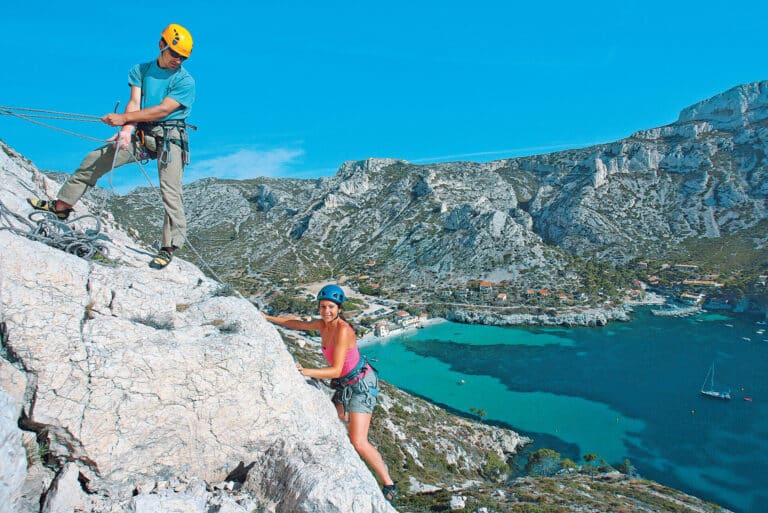 To personer klatrer på en solskinsdag med udsigt over et naturskønt kystlandskab med turkisblåt vand og klipper. Klatrerne bærer hjelme og klatreudstyr, og baggrunden omfatter en bugt med både og en lille bebyggelse.