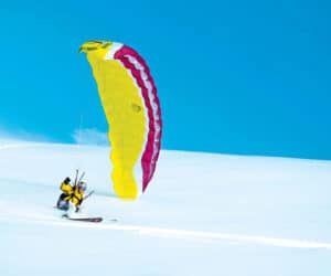 En person deltager i snowkiting over et snedækket landskab ved at bruge en stor gul og pink drage, mens han står på ski. De er klædt i vinterudstyr under en klar blå himmel, hvor sneen sparker op under dem, mens de glider hen over sneen.