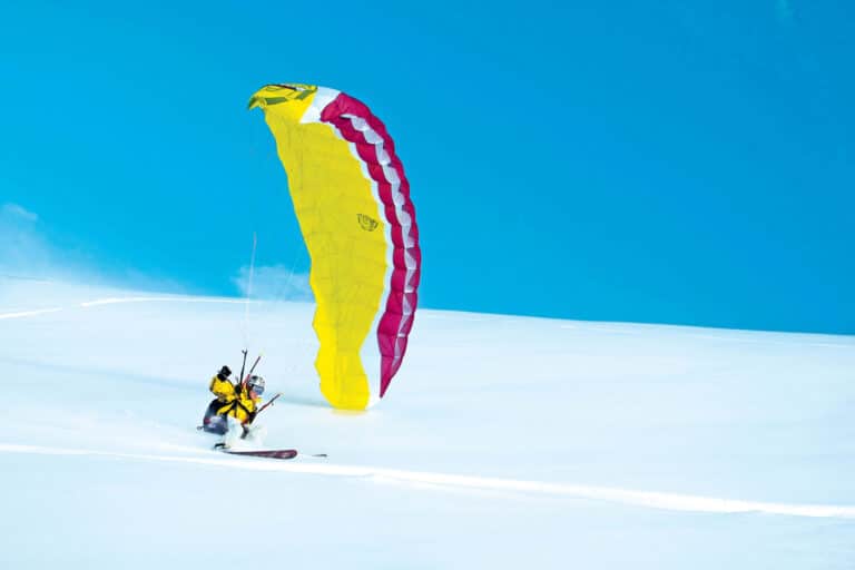 En person deltager i snowkiting over et snedækket landskab ved at bruge en stor gul og pink drage, mens han står på ski. De er klædt i vinterudstyr under en klar blå himmel, hvor sneen sparker op under dem, mens de glider hen over sneen.