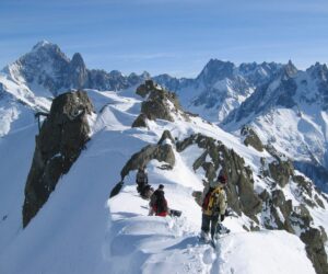 En gruppe klatrere med gear vandrer langs en sneklædt bjergryg, omgivet af forrevne tinder under en klar blå himmel. Majestætiske bjerge danner baggrunden og skaber en betagende alpine scene.