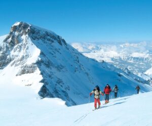 En gruppe mennesker i vinterudstyr vandrer op ad en sneklædt bjergskråning under en klar blå himmel. En robust, snedækket top er tydeligt synlig til venstre med en stor bjergkæde i baggrunden.