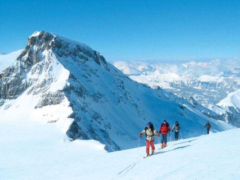 En gruppe mennesker i vinterudstyr vandrer op ad en sneklædt bjergskråning under en klar blå himmel. En robust, snedækket top er tydeligt synlig til venstre med en stor bjergkæde i baggrunden.
