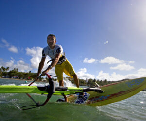 En person, der windsurferer energisk hen over vandet under en klar blå himmel. De er iført en hvid top og gule shorts, med frodige grønne træer og en fjern kystlinje i baggrunden.