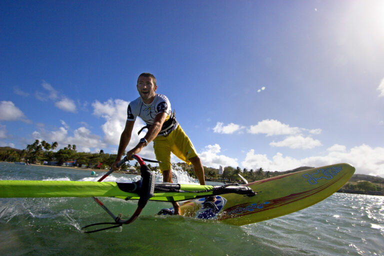 En person, der windsurferer energisk hen over vandet under en klar blå himmel. De er iført en hvid top og gule shorts, med frodige grønne træer og en fjern kystlinje i baggrunden.