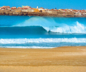 En naturskøn udsigt over havets bølger, der slår ned på en sandstrand med en kystby og klippeklipper i baggrunden under en klar blå himmel.