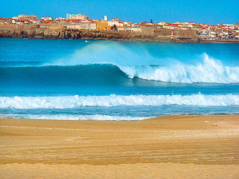 En naturskøn udsigt over havets bølger, der slår ned på en sandstrand med en kystby og klippeklipper i baggrunden under en klar blå himmel.