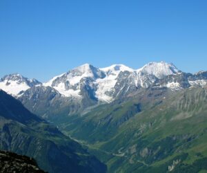 Sneklædte bjerge under en klar blå himmel med frodige grønne dale og stenede skråninger i forgrunden. Barske tinder strækker sig over horisonten og skaber et malerisk alpelandskab.