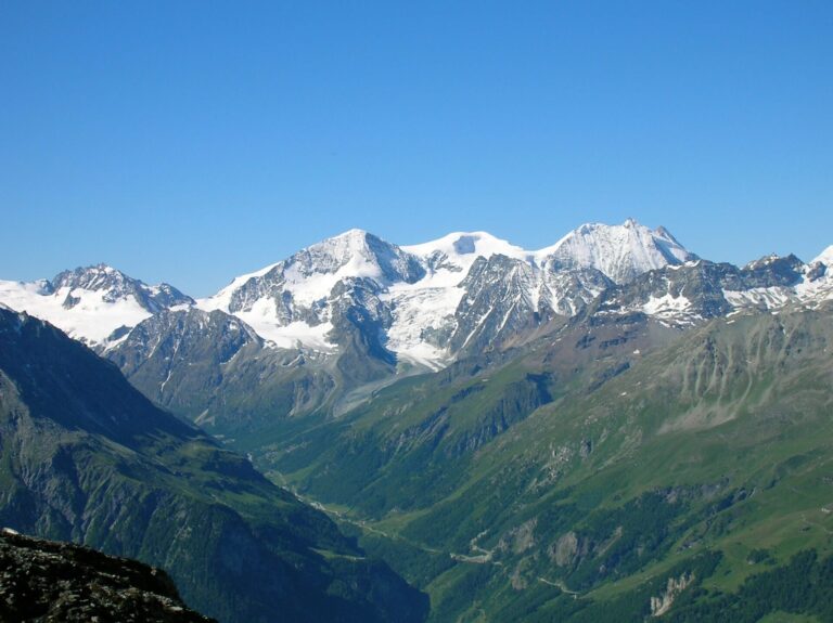Sneklædte bjerge under en klar blå himmel med frodige grønne dale og stenede skråninger i forgrunden. Barske tinder strækker sig over horisonten og skaber et malerisk alpelandskab.
