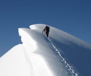 En enlig bjergbestiger bestiger en snedækket højderyg under en klar blå himmel. Stien er smal og omgivet af stejle skråninger, hvilket fremhæver den udfordrende stigning. Klatreren bærer udstyr og bærer hjelm, hvilket understreger eventyret i fjerntliggende omgivelser.