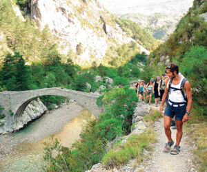 En gruppe vandrere går langs en smal sti med udsigt over en stenbuebro og en flod nedenfor. Scenen er omgivet af frodigt grønt og bjergrigt terræn under en klar himmel.
