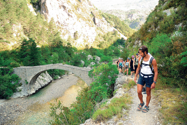 En gruppe vandrere går langs en smal sti med udsigt over en stenbuebro og en flod nedenfor. Scenen er omgivet af frodigt grønt og bjergrigt terræn under en klar himmel.