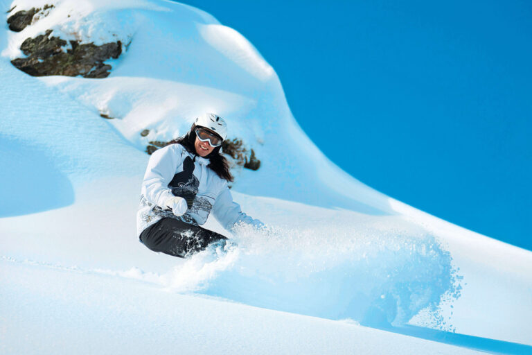 En person klædt i vinterudstyr, inklusive en hvid hjelm og beskyttelsesbriller, snowboards ned ad en sneklædt skråning under en klar blå himmel, og sparker en spray af sne op bag sig. Snedækkede sten er synlige i baggrunden.