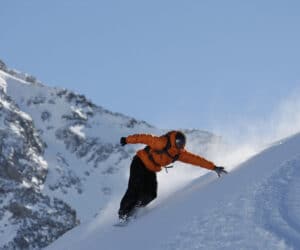 En snowboarder i orange jakke og sorte bukser skærer ned ad en sneklædt bjergskråning mod en klar blå himmel. Sne sprøjter op, mens de laver et skarpt sving, med forrevne, snedækkede toppe i baggrunden.