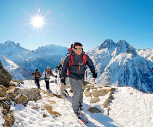 En gruppe mennesker iført rygsække og udstyr vandrer hen over et snedækket bjerglandskab under en skarp sol. Barske tinder og klar blå himmel danner baggrunden.