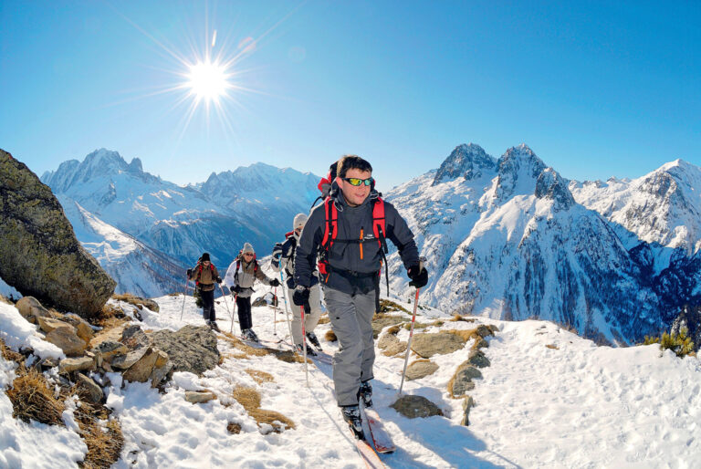 En gruppe mennesker iført rygsække og udstyr vandrer hen over et snedækket bjerglandskab under en skarp sol. Barske tinder og klar blå himmel danner baggrunden.