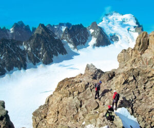 Vandrere i røde jakker navigerer i klippefyldt terræn i et bjergrigt landskab med snedækkede tinder og en klar blå himmel.