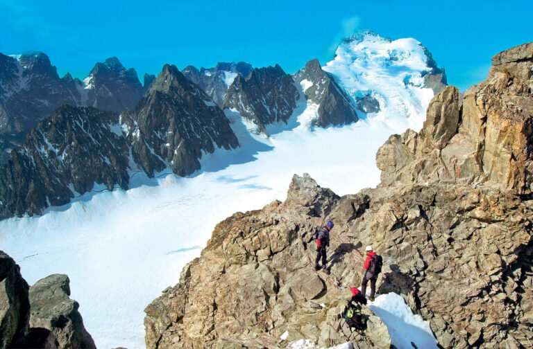 Vandrere i røde jakker navigerer i klippefyldt terræn i et bjergrigt landskab med snedækkede tinder og en klar blå himmel.