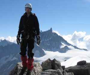 En person iført klatreudstyr står på en klippet bjergtop med sneklædte bjerge og en klar blå himmel i baggrunden.