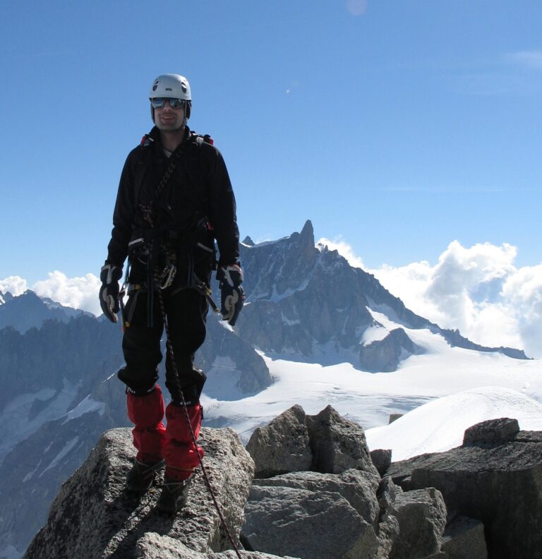 En person iført klatreudstyr står på en klippet bjergtop med sneklædte bjerge og en klar blå himmel i baggrunden.