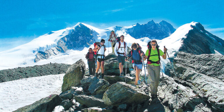 En gruppe vandrere med rygsække og trekkingstave krydser stenet terræn i et bjergrigt område. Sneklædte tinder og en klar blå himmel giver en fantastisk kulisse.