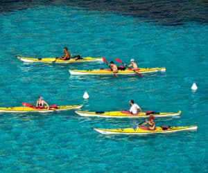 En gruppe mennesker, der sejler i kajak på klart blåt vand med hvide bøjer omkring sig. Kajakkerne er gule, og hver person padler med røde årer. Scenen er levende og afspejler en solskinsdag.