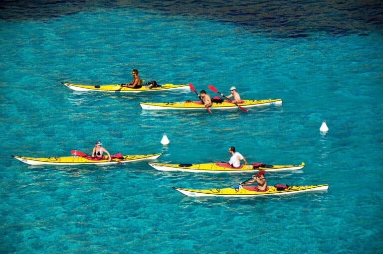 En gruppe mennesker, der sejler i kajak på klart blåt vand med hvide bøjer omkring sig. Kajakkerne er gule, og hver person padler med røde årer. Scenen er levende og afspejler en solskinsdag.