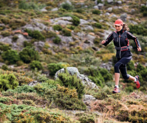 En person i atletisk gear løber gennem et klippefyldt, bakket landskab. De bærer kasket, jakke og sneakers. Baggrunden er fyldt med buske og ujævnt terræn.