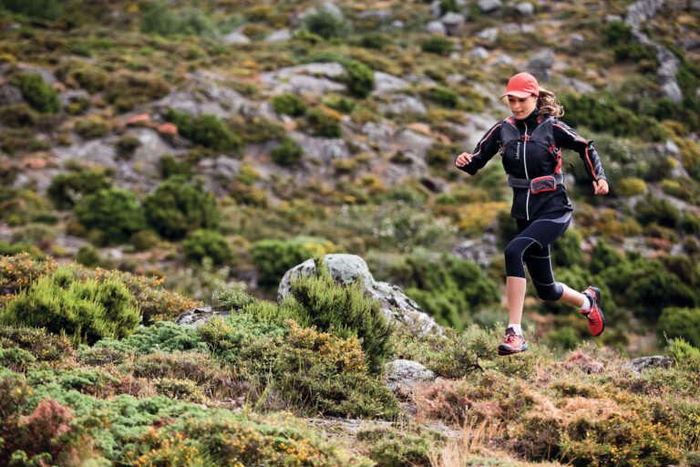 En person i atletisk gear løber gennem et klippefyldt, bakket landskab. De bærer kasket, jakke og sneakers. Baggrunden er fyldt med buske og ujævnt terræn.