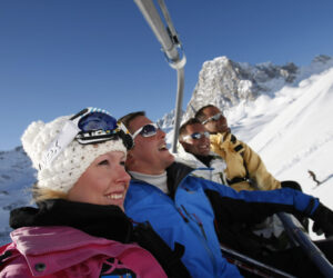 En gruppe på fire personer i vintertøj kører i en skilift på en solskinsdag. De smiler og er iført skiudstyr på baggrund af snedækkede bjerge og en klar blå himmel.