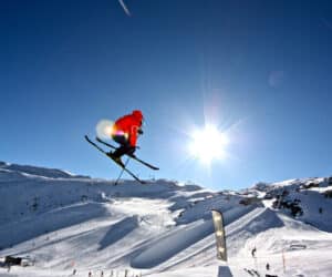En skiløber i et knaldrødt outfit udfører et spring i luften mod en klar blå himmel. Snedækkede bjerge og skiløjper er synlige i baggrunden under en skarp sol.