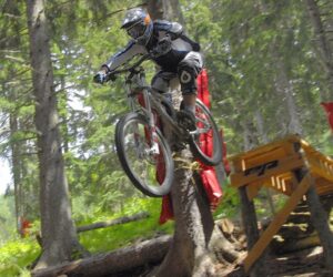 En mountainbiker iført beskyttelsesudstyr svæver gennem luften og hopper fra en trærampe i skoven. Det er et spændende sportsferieeventyr med frodige omgivelser, høje træer og sollys. Rød polstring er viklet rundt om nogle træstammer for sikkerheden.