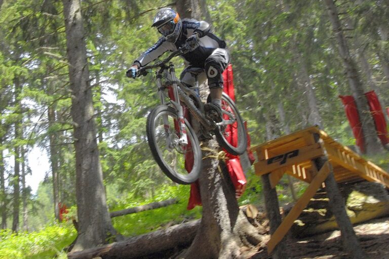 En mountainbiker iført beskyttelsesudstyr svæver gennem luften og hopper fra en trærampe i skoven. Det er et spændende sportsferieeventyr med frodige omgivelser, høje træer og sollys. Rød polstring er viklet rundt om nogle træstammer for sikkerheden.