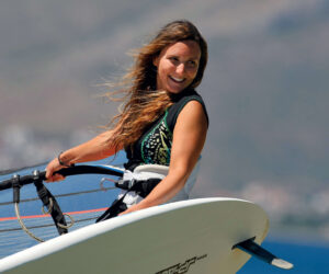 En kvinde med langt hår, smilende, holder et windsurfingbræt. Hun er iført en sort og hvid top, stående mod en sløret naturlig baggrund med bakker og en klar himmel.
