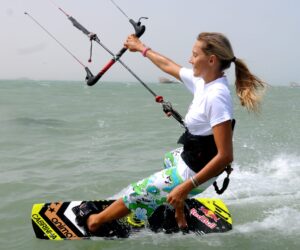 En kvinde kiteboarder på vandet og holder fast i kontrolstangen med begge hænder. Hun er iført en hvid skjorte og blomstermønstrede boardshorts og glider over bølgerne med et fokuseret udtryk.