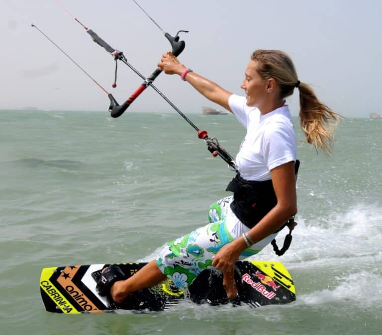 En kvinde kiteboarder på vandet og holder fast i kontrolstangen med begge hænder. Hun er iført en hvid skjorte og blomstermønstrede boardshorts og glider over bølgerne med et fokuseret udtryk.