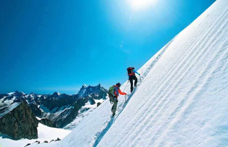 To klatrere bestiger en stejl, sneklædt bjergskråning under en klar blå himmel. Skarvede tinder omgiver dem i baggrunden, og solen skinner klart og skaber en dramatisk alpescene.