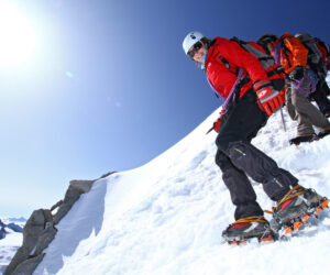 To klatrere iført hjelme og rygsække navigerer på en sneklædt bjergskråning under en lys, klar himmel. Klatreren foran bærer en rød jakke og er placeret lavere på skråningen, vinklet lidt nedad. Robuste tinder er synlige i baggrunden.