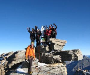En gruppe klatrere, udstyret med hjelme og udstyr, står triumferende på en klippet bjergtop under en klar blå himmel. En klatrer i en orange jakke står lidt fra hinanden i forgrunden. Snepletter er synlige på klipperne.
