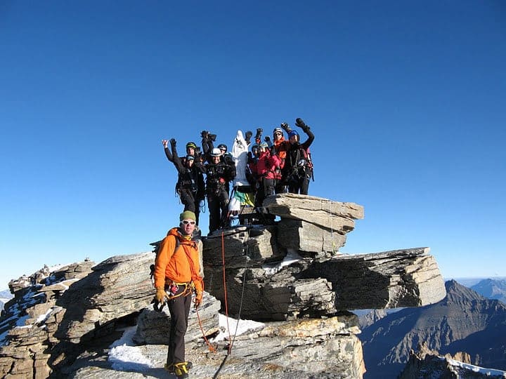 En gruppe klatrere, udstyret med hjelme og udstyr, står triumferende på en klippet bjergtop under en klar blå himmel. En klatrer i en orange jakke står lidt fra hinanden i forgrunden. Snepletter er synlige på klipperne.