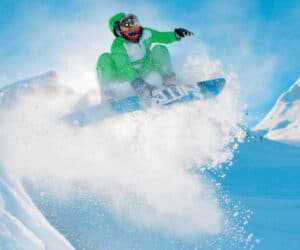 En snowboarder i et levende grønt outfit udfører et lufttrick mod en klar blå himmel. Sne sprøjter rundt, mens de manøvrerer gennem et sneklædt bjerglandskab og fanger et dynamisk og energisk øjeblik.