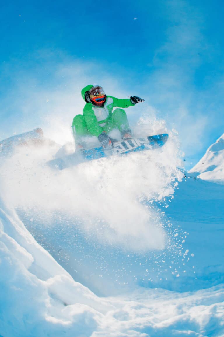 En snowboarder i et levende grønt outfit udfører et lufttrick mod en klar blå himmel. Sne sprøjter rundt, mens de manøvrerer gennem et sneklædt bjerglandskab og fanger et dynamisk og energisk øjeblik.