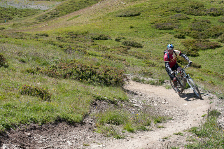En mountainbiker i rød trøje og hjelm kører ned ad en snoet jordsti gennem frodige grønne bakker med pletter af buske og græs på en solskinsdag.