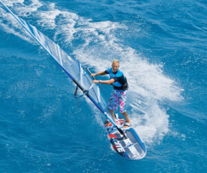 En person, der windsurfer på klart blåt vand, balancerer på et bræt, mens han holder fast i et sejl. Personen er iført en sort og blå våddragt og farverige shorts, hvilket skaber en livlig scene på baggrund af det pulserende hav.